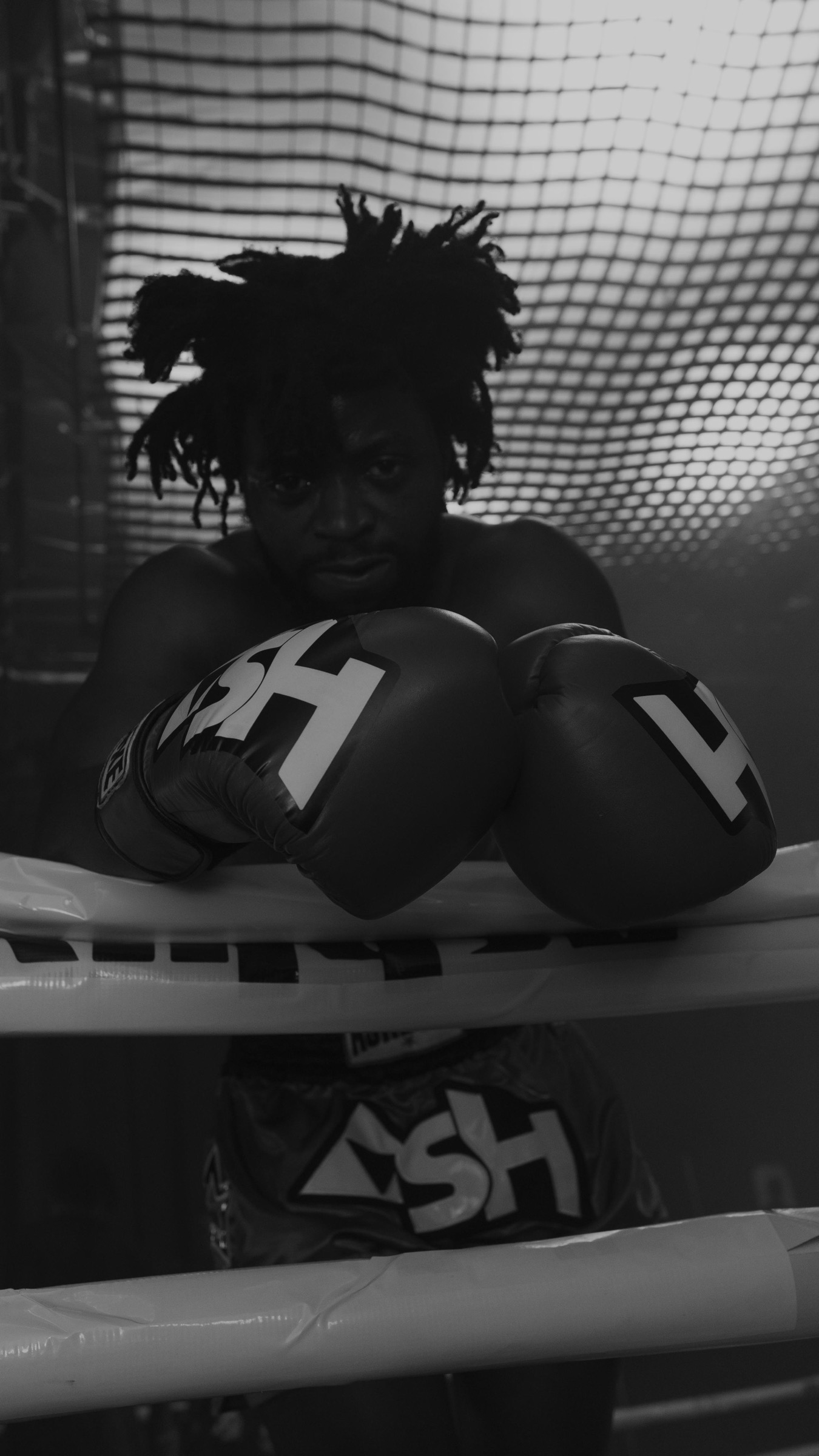 Black and white portrait of a boxer leaning on the ropes, gloves crossed, looking directly at camera.